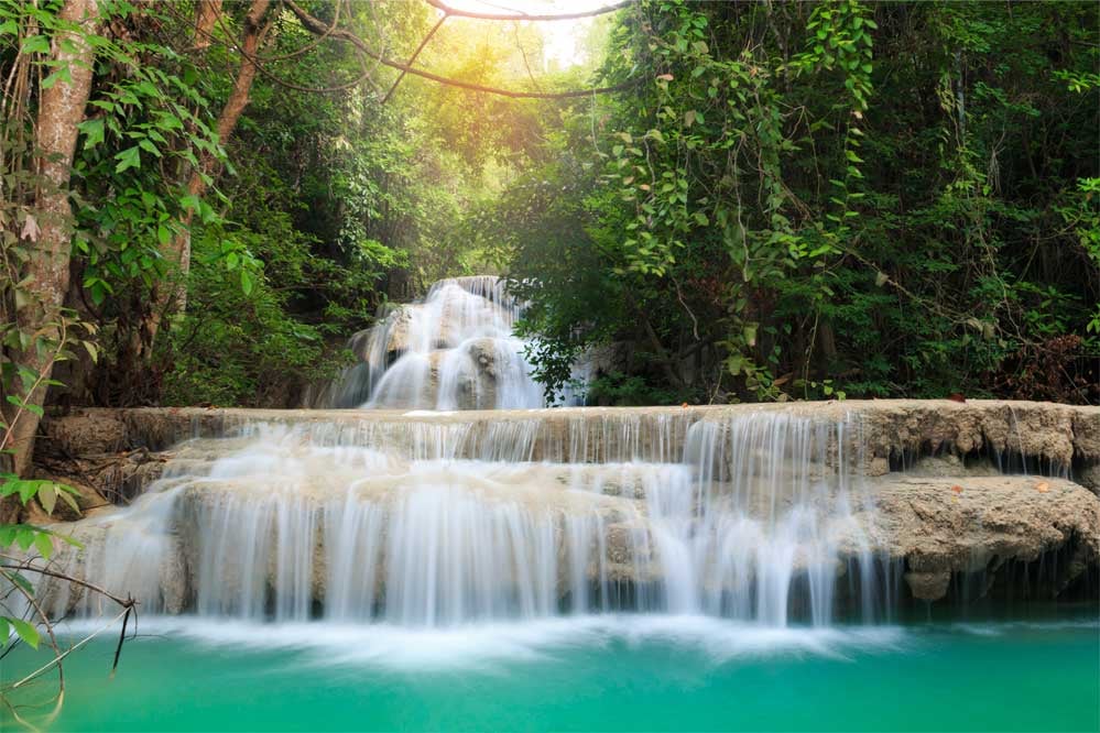 Chute d'eau dans la jungle avec rayons de soleil filtrant à travers les arbres, dans le par d'Erawan en Thaïlande.